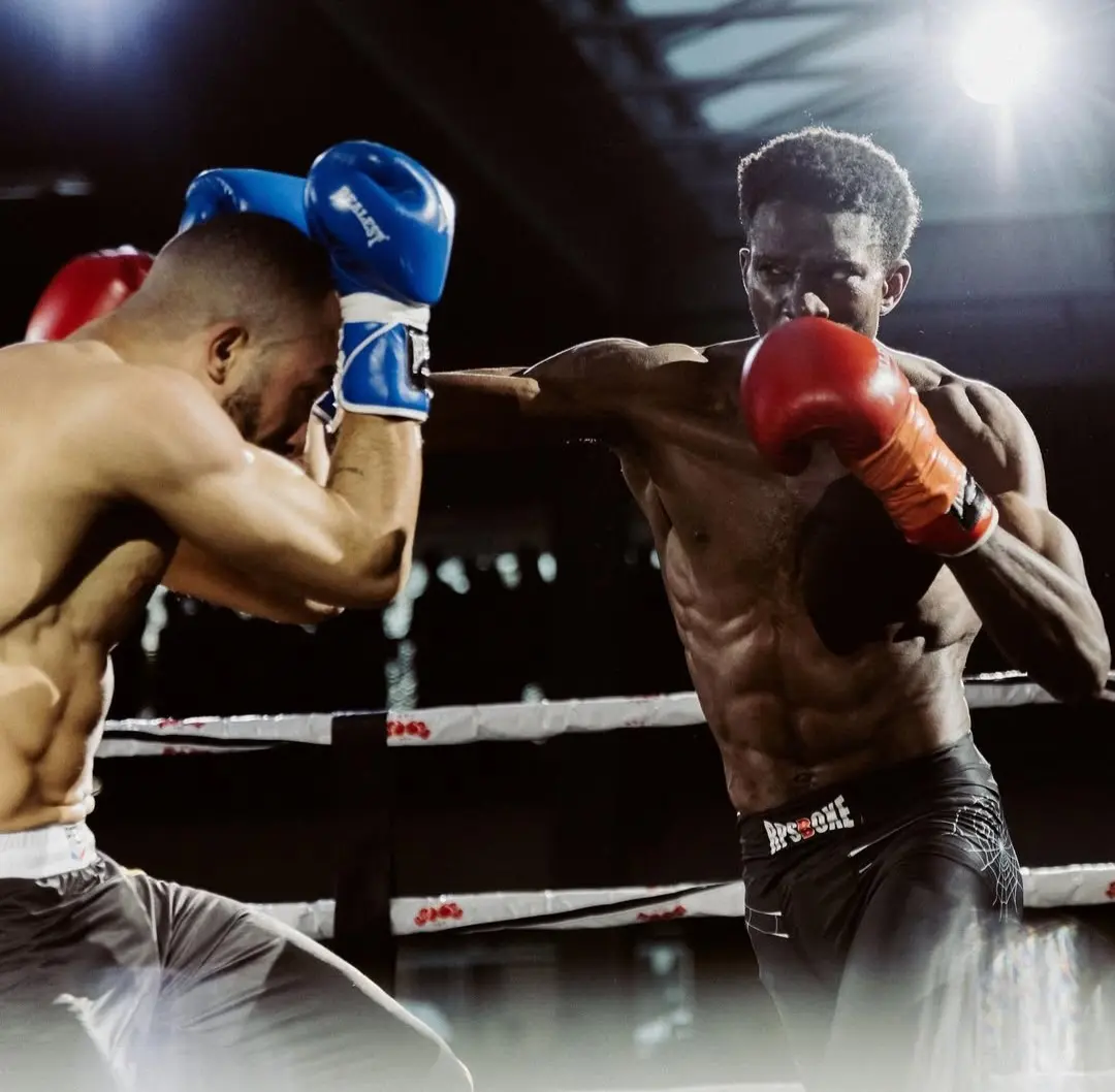 Deux boxeurs en combat dans le ring: l’un en bleu bloque un coup de l’autre en rouge; muscles, gants et ambiance de salle.