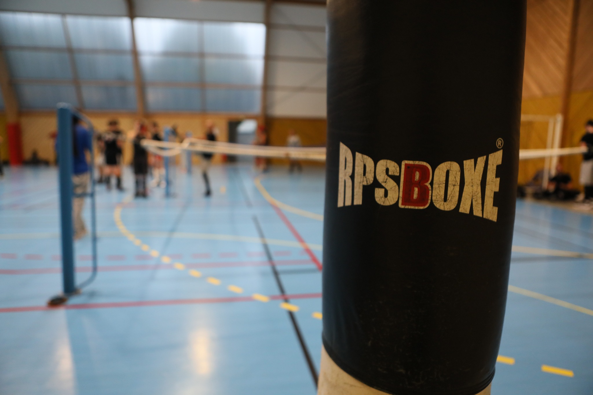 Gant de boxe compétition posé sur un sac RPSBOXE dans une salle d’entraînement avec des personnes et un ring en arrière-plan.